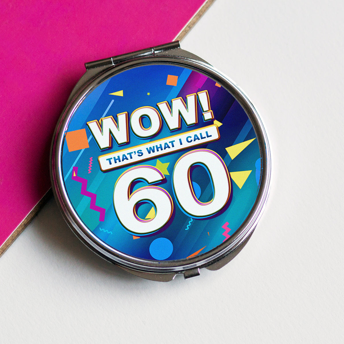 A round compact mirror with a colorful retro 1980s-style design featuring the text 'WOW! THAT'S WHAT I CALL 60' in bold white letters. The compact is positioned diagonally on a split background of bright pink and off-white.