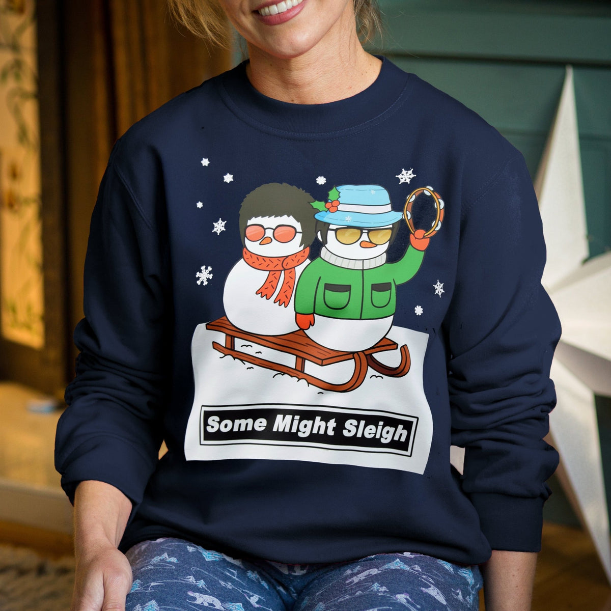 Person wearing a navy blue Christmas jumper with a graphic of Liam and Noel. Gallagher as two snowmen riding a sledge and text 'Some Might Sleigh', sitting indoors.