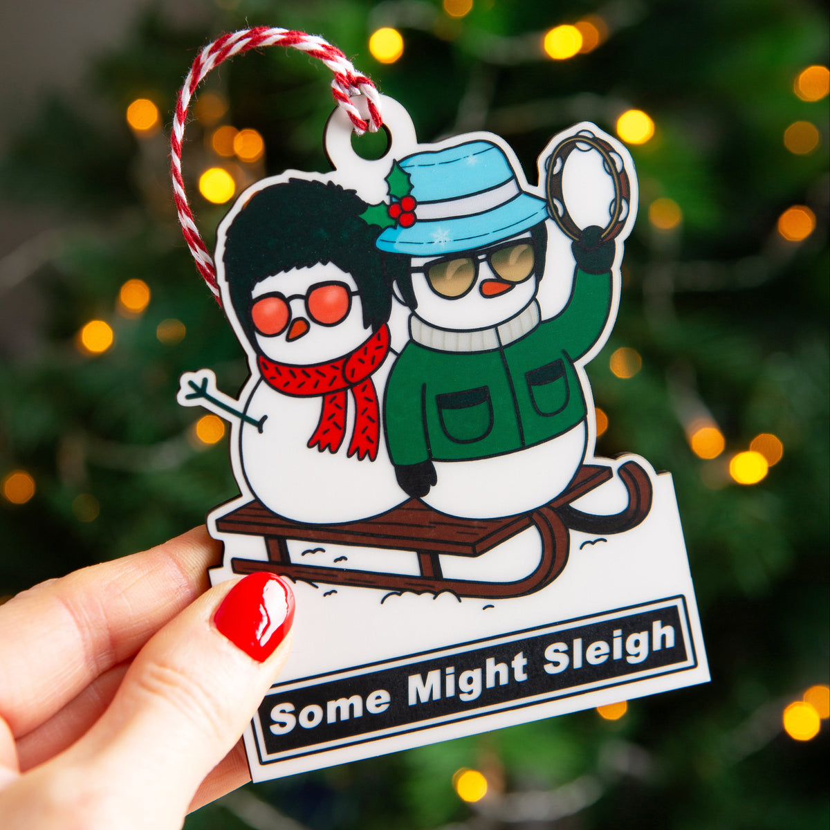 A hand with red nail polish holds a die-cut Christmas ornament featuring Liam and Noel on a sleigh, with a red-and-white hanging loop and the text 'Some Might Sleigh' at the bottom. The background is a blurred Christmas tree with warm yellow lights.