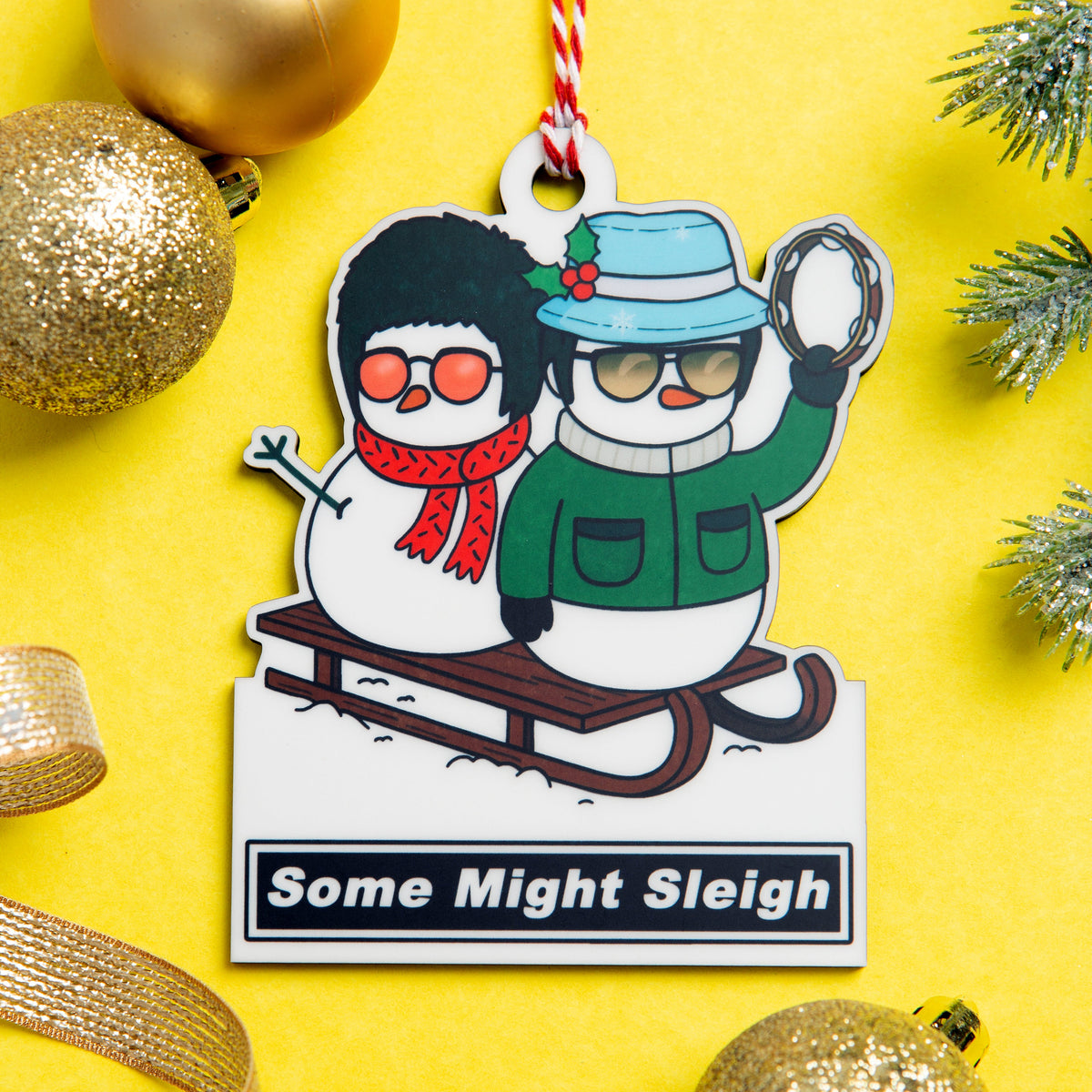 A cartoon Liam and Noel Gallagher on a sled hanging ornament with the text 'Some Might Sleigh' on a bright yellow background with holiday decorations.