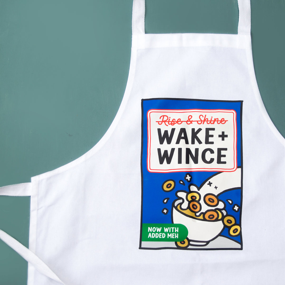 A white apron with a colorful cereal box graphic that reads 'Rise & Shine WAKE + WINCE' and 'NOW WITH ADDED MEH', shown against a muted teal background.