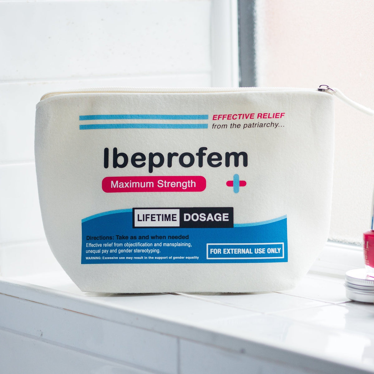 A white plastic zippered pouch of Ibuprofen Maximum Strength with blue and pink text and graphics, sitting on a white tiled bathroom counter next to a red bottle.