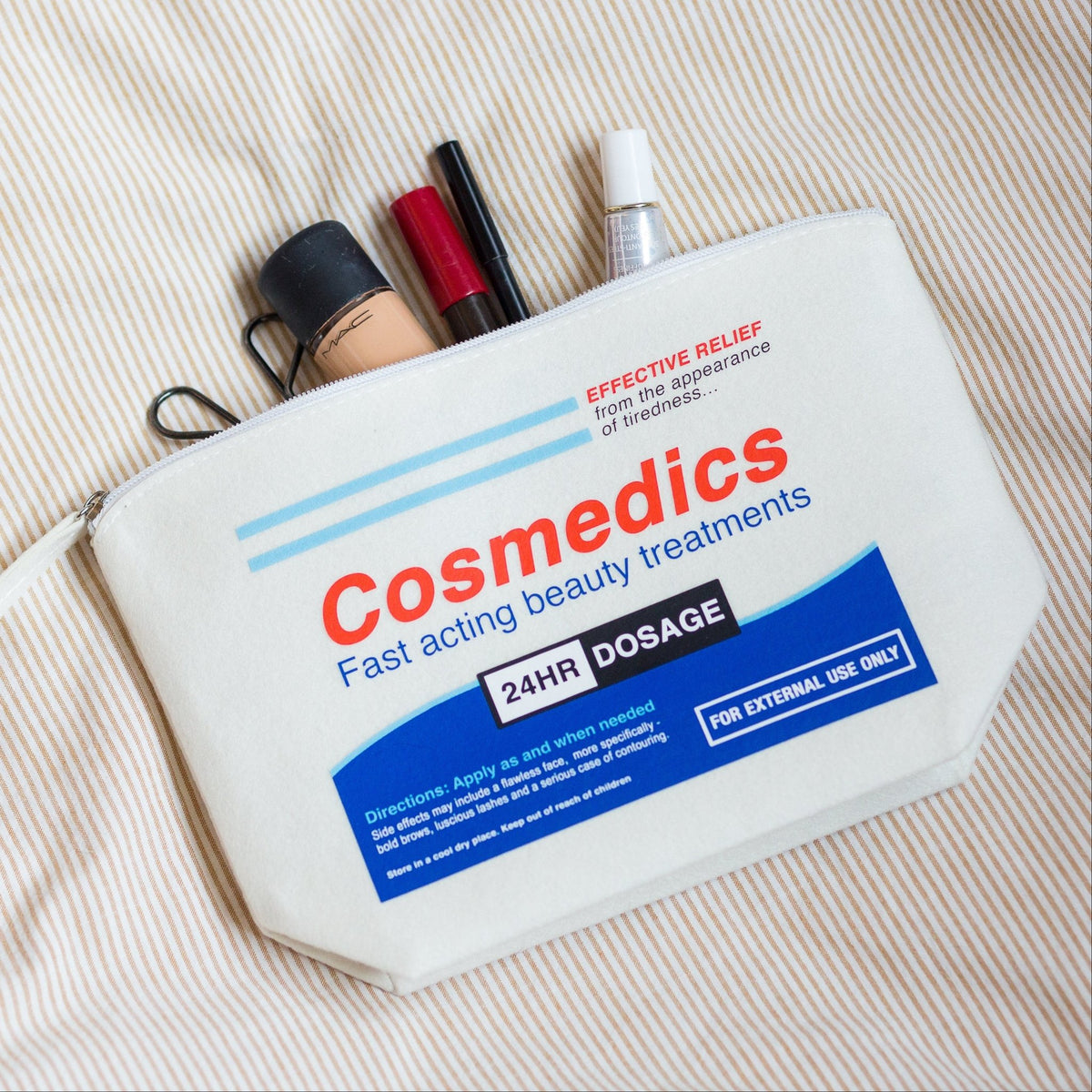 A white cosmetic pouch printed with 'Cosmedics Fast acting beauty treatments' in red and blue text, shown on a beige striped fabric background with makeup items partially sticking out of the top.