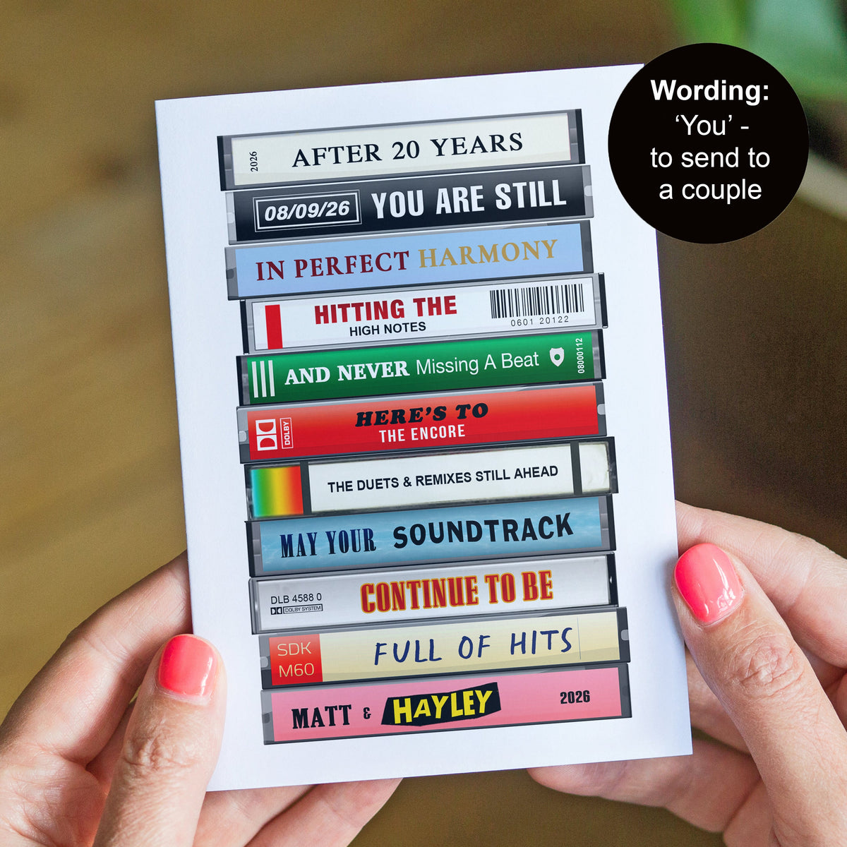 A person holds a printed card featuring stacked cassette tape graphics with song titles and romantic phrases. A black circular sticker reads 'Wording: 'You' - to send to a couple.