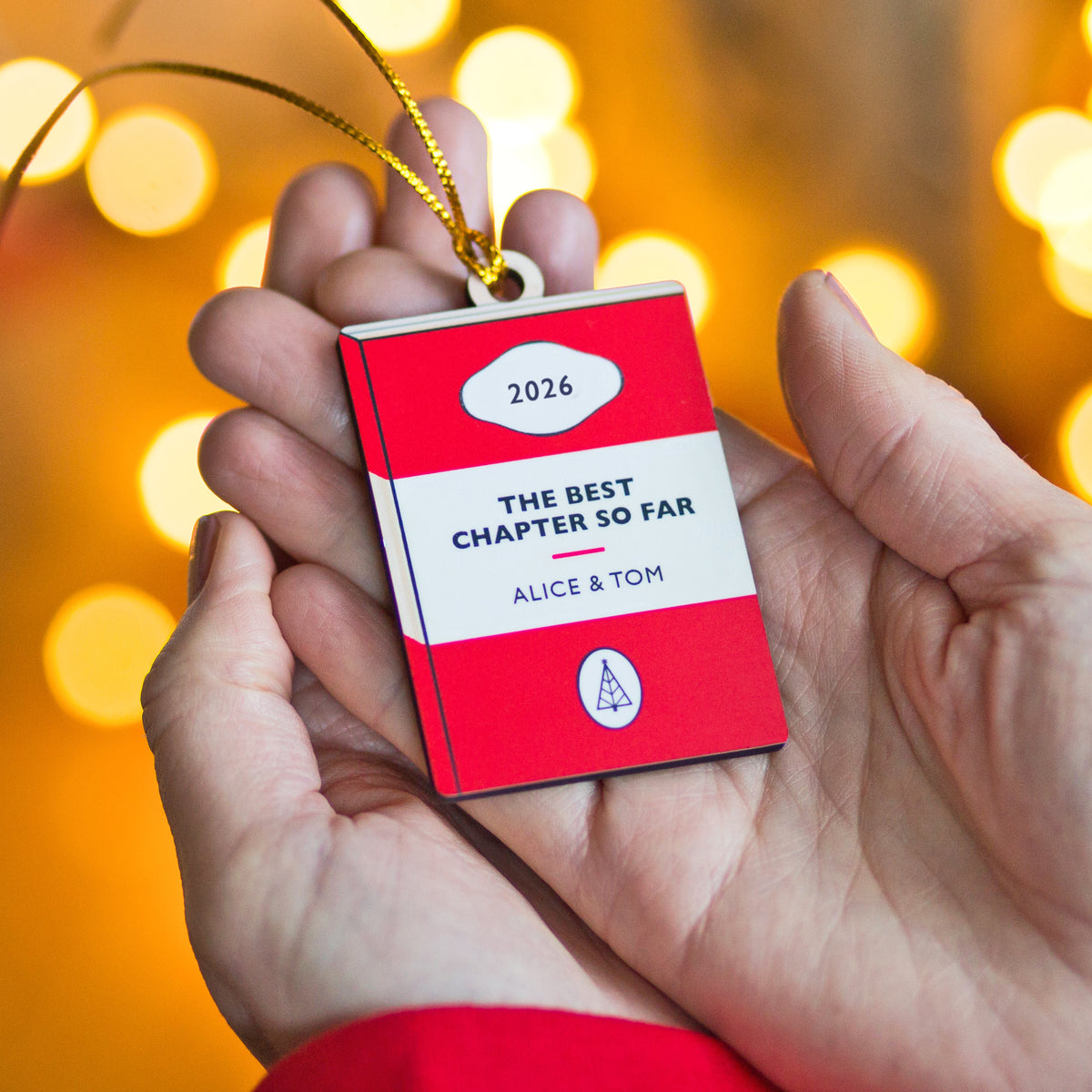 A person's hands holding a red and white book-shaped ornament with a gold hanging string. The ornament reads '2026', 'THE BEST CHAPTER SO FAR', and 'ALICE & TOM'.