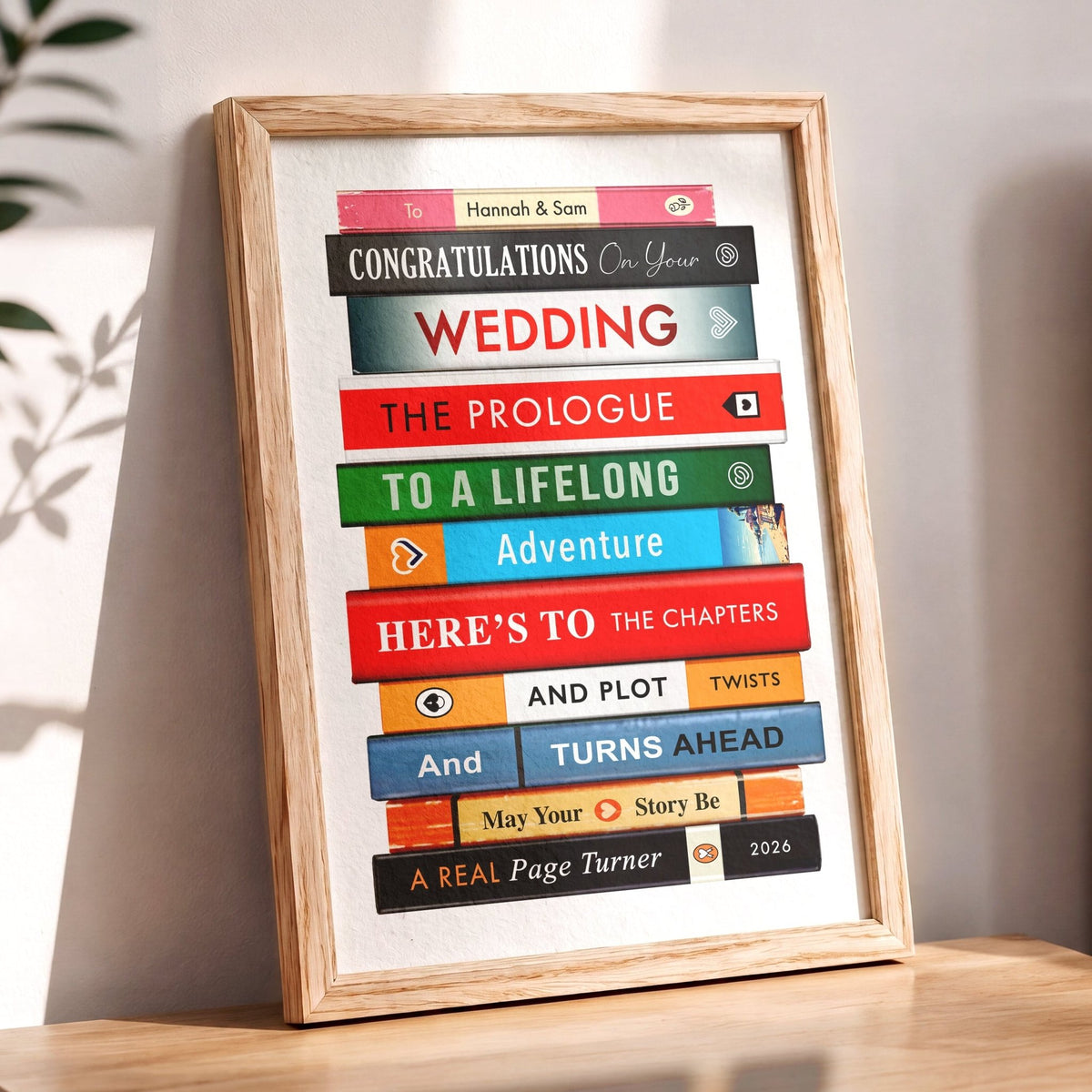 A personalised framed book-stack art print for a wedding, leaning on a wooden table against a light wall. The print features colorful book spines stacked to form sentences like 'Congratulations on your WEDDING' and 'The Prologue to a Lifelong Adventure'.