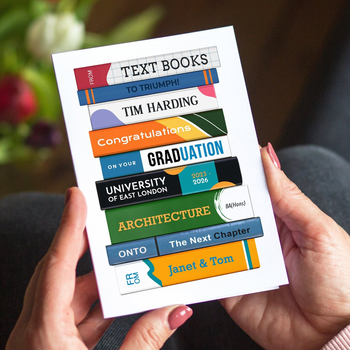 Graduation card with colorful text books design held by hands, with flowers in the background.