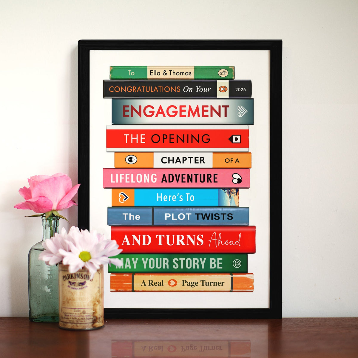 A framed personalised book cover art print displayed on a wooden table next to a glass vase with pink flowers. The print features colorful book spines stacked to form a story outline with text such as 'To Ella & Thomas', 'CONGRATULATIONS On Your', and 'ENGAGEMENT'.