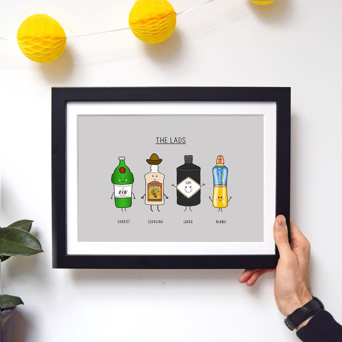 Framed print with cartoon bottles labeled 'The Lads' held by a hand on a white background.