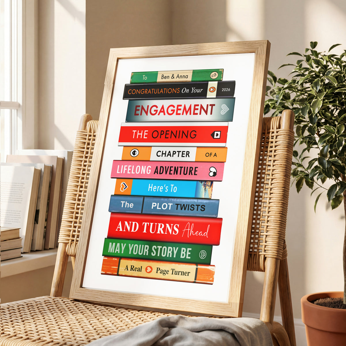 A framed personalised book cover art print leaning on a woven chair in a bright room. The print features stacked colorful book spines with text like 'ENGAGEMENT', 'THE OPENING', and 'LIFELONG ADVENTURE'.