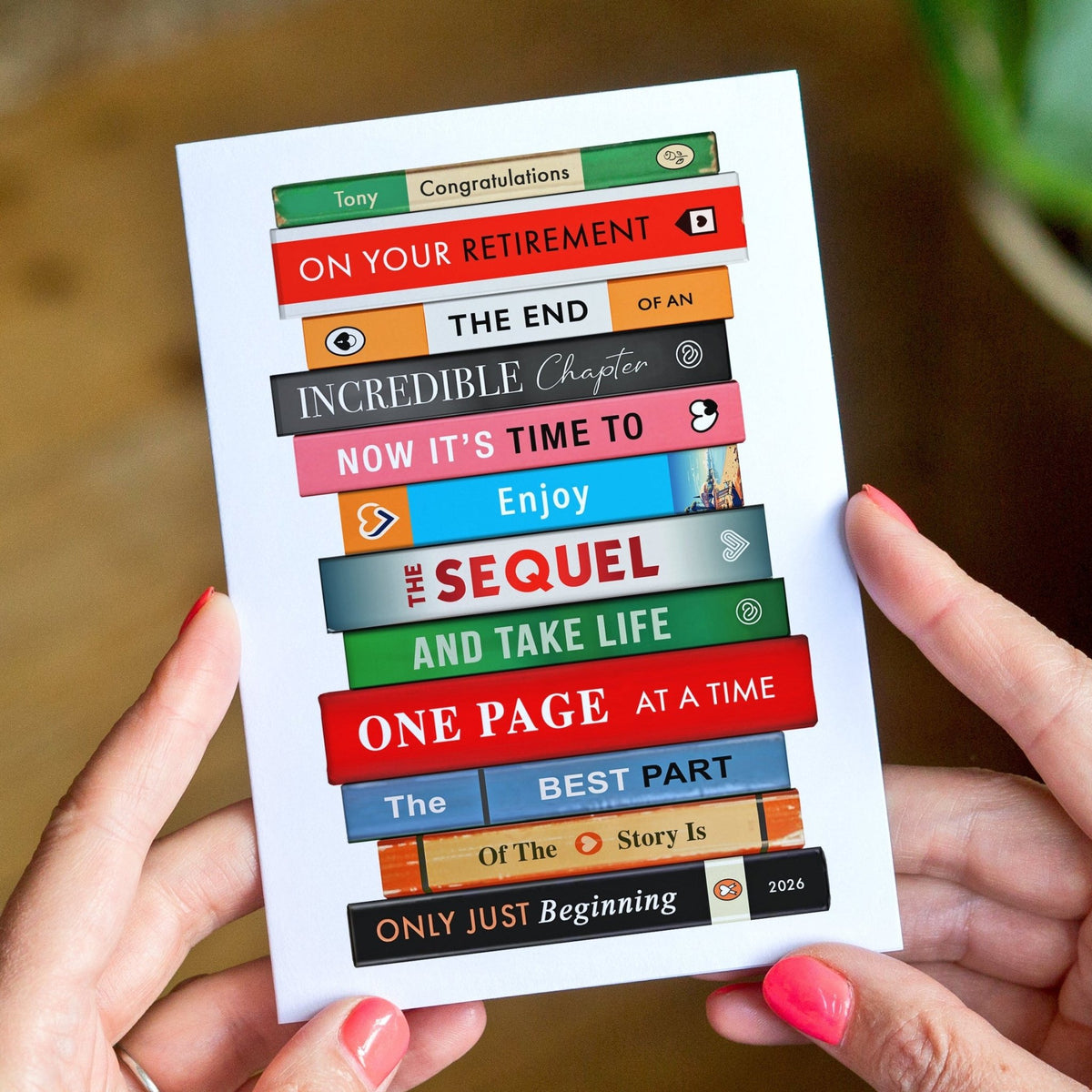 A person holds a white card featuring colorful book spine graphics with retirement-themed text such as 'Congratulations,' 'The End,' 'The Sequel,' and 'One Page at a Time.' The background is a softly blurred indoor setting with warm wood tones and a hint of greenery.