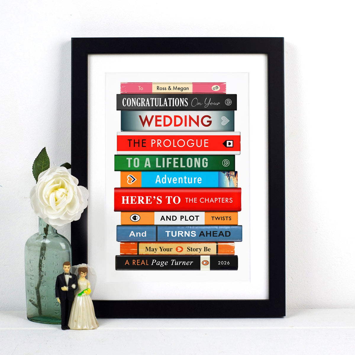 A framed personalised book-stack wedding art print displayed on a white surface next to a small vase with a white rose and a miniature wedding figurine. The print features colorful book spines with wedding-themed text like 'Congratulations on your Wedding' and 'The Prologue'.