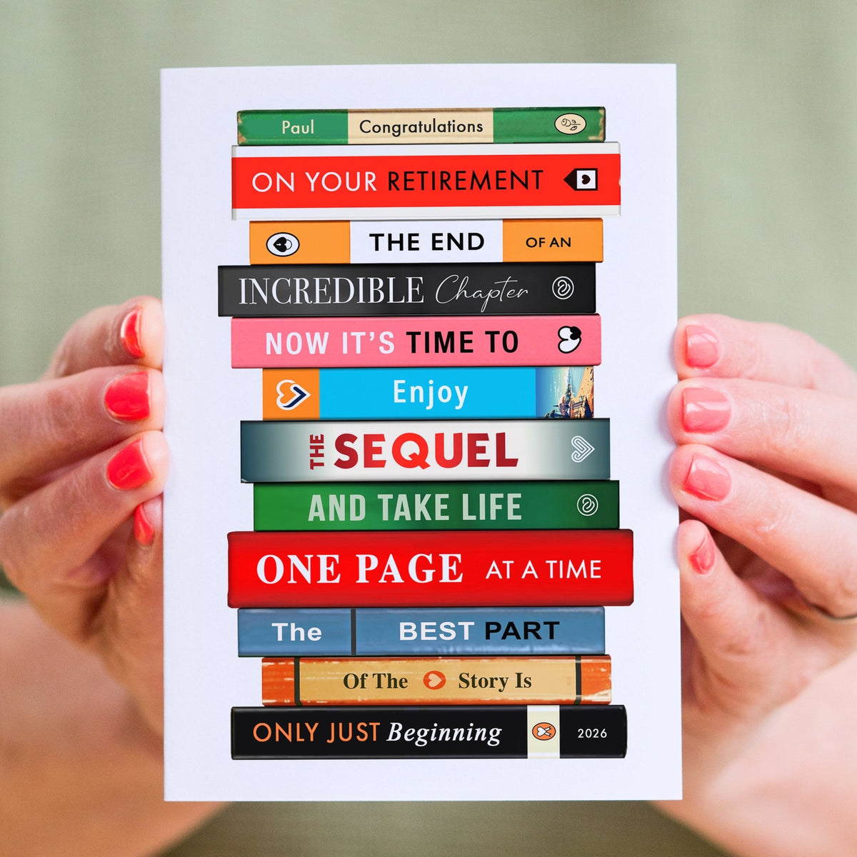 A white greeting card held by two hands with pink manicured nails. The card features a stack of colorful book spines with humorous retirement-themed text such as 'ON YOUR RETIREMENT', 'THE END OF AN INCREDIBLE Chapter', and 'ONE PAGE AT A TIME'.
