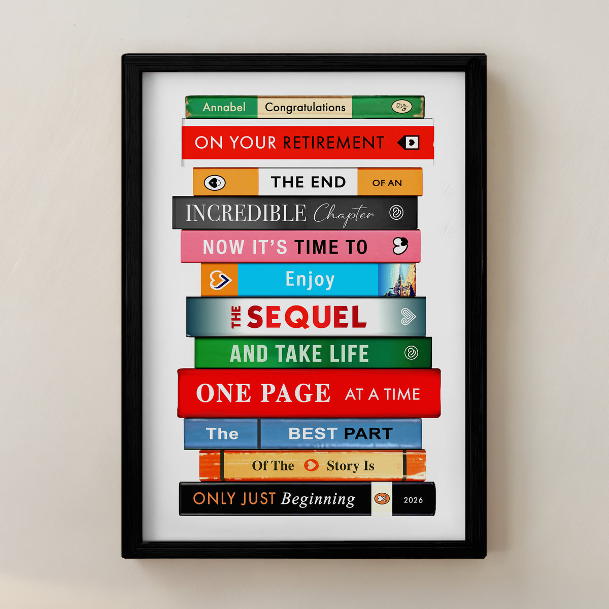 A framed personalized book spine collage print with colorful book spines and text messages, enclosed in a black frame against a light beige wall.