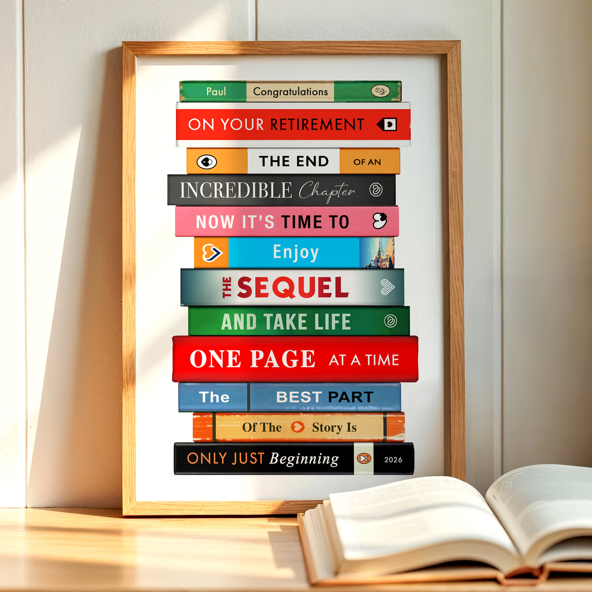 A framed personalized book-stack retirement poster leaning against a light wood-paneled wall on a wooden surface, with an open book in the foreground. The poster features colorful book-spine graphics with motivational retirement phrases like 'ON YOUR RETIREMENT' and 'ONE PAGE AT A TIME'.