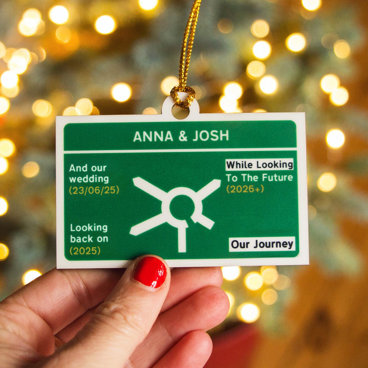 Hand holding a green road sign christmas tree ornament with text 'ANNA & JOSH' against a blurred festive background.