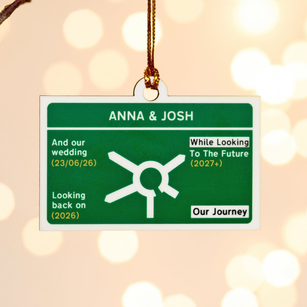 A rectangular Christmas tree ornament with a green and white design, hanging from a gold string against a warm, blurred bokeh background. The ornament features personalised text for 'ANNA & JOSH' and important milestones. The ornament is designed to look like a UK road sign