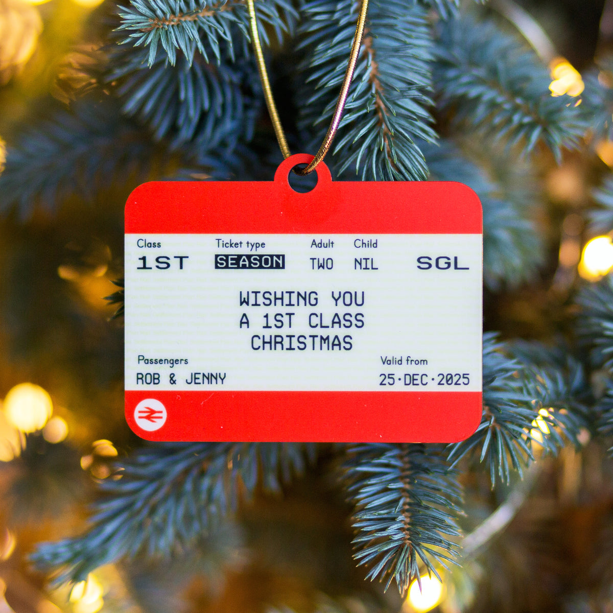 An orange and white Christmas ornament designed to look like a train ticket, hanging from a gold string on a pine tree with warm bokeh lights in the background. The ticket has personalized text reading 'WISHING YOU A 1ST CLASS CHRISTMAS' and passenger names 'ROB & JENNY'.