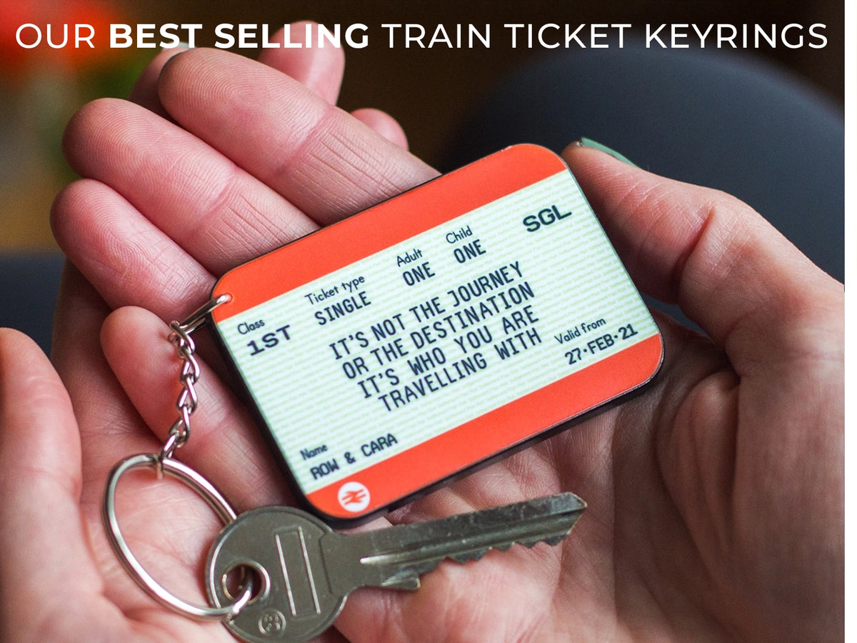 A person's hands holding a novelty train ticket keyring with a metal key attached. The keyring is shaped like an orange and white train ticket with printed text and a silver split ring and key.