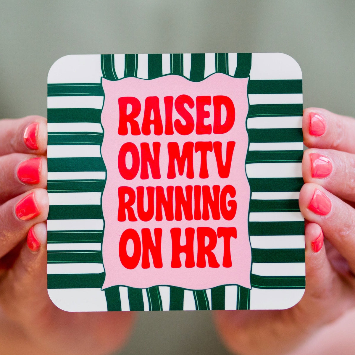 Coaster with text 'Raised on MTV Running on HRT' held by hands with red nail polish.