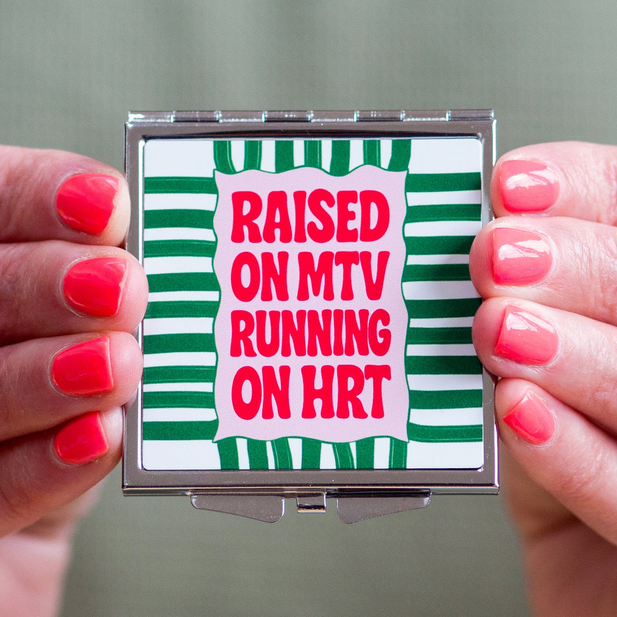 Compact mirror with 'Raised on MTV Running on HRT' text held by hands with red nail polish.