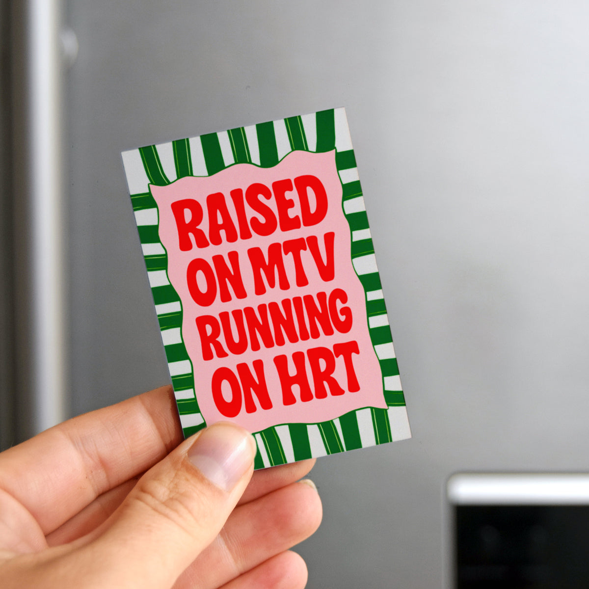 Hand holding a fridge magnet with text 'Raised on MTV Running on HRT' against a refrigerator