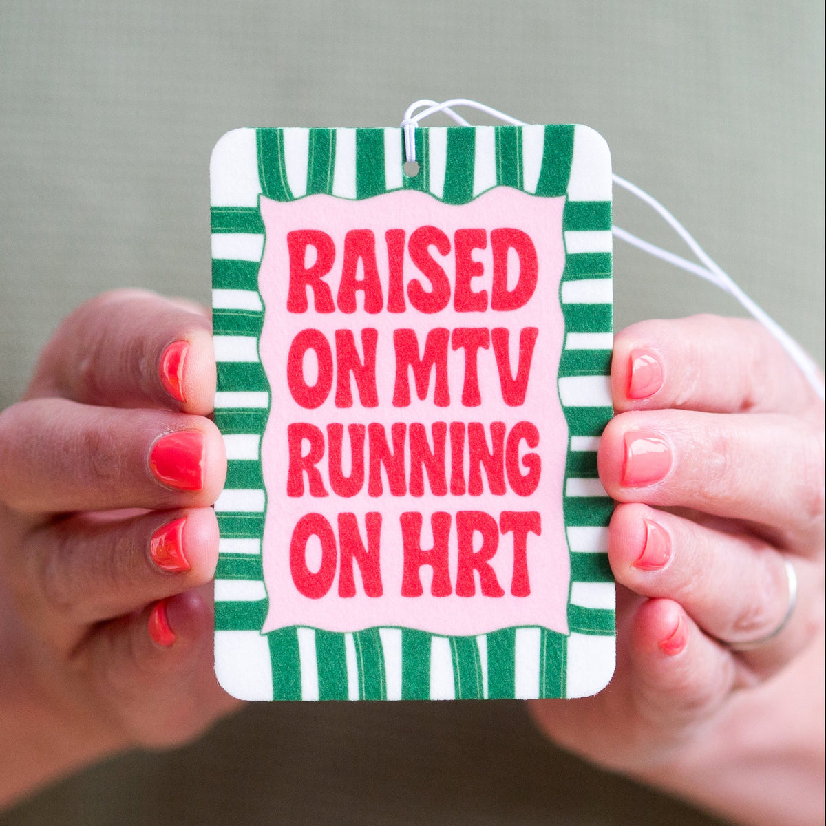 Hand holding a striped car air freshener with text 'Raised on MTV Running on HRT' against a neutral background