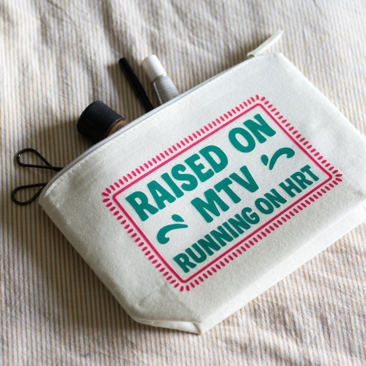 An off-white canvas zippered pouch with a printed graphic reading 'RAISED ON MTV RUNNING ON HRT' in teal and pink, shown on a textured beige fabric background with small accessories peeking out from the top.