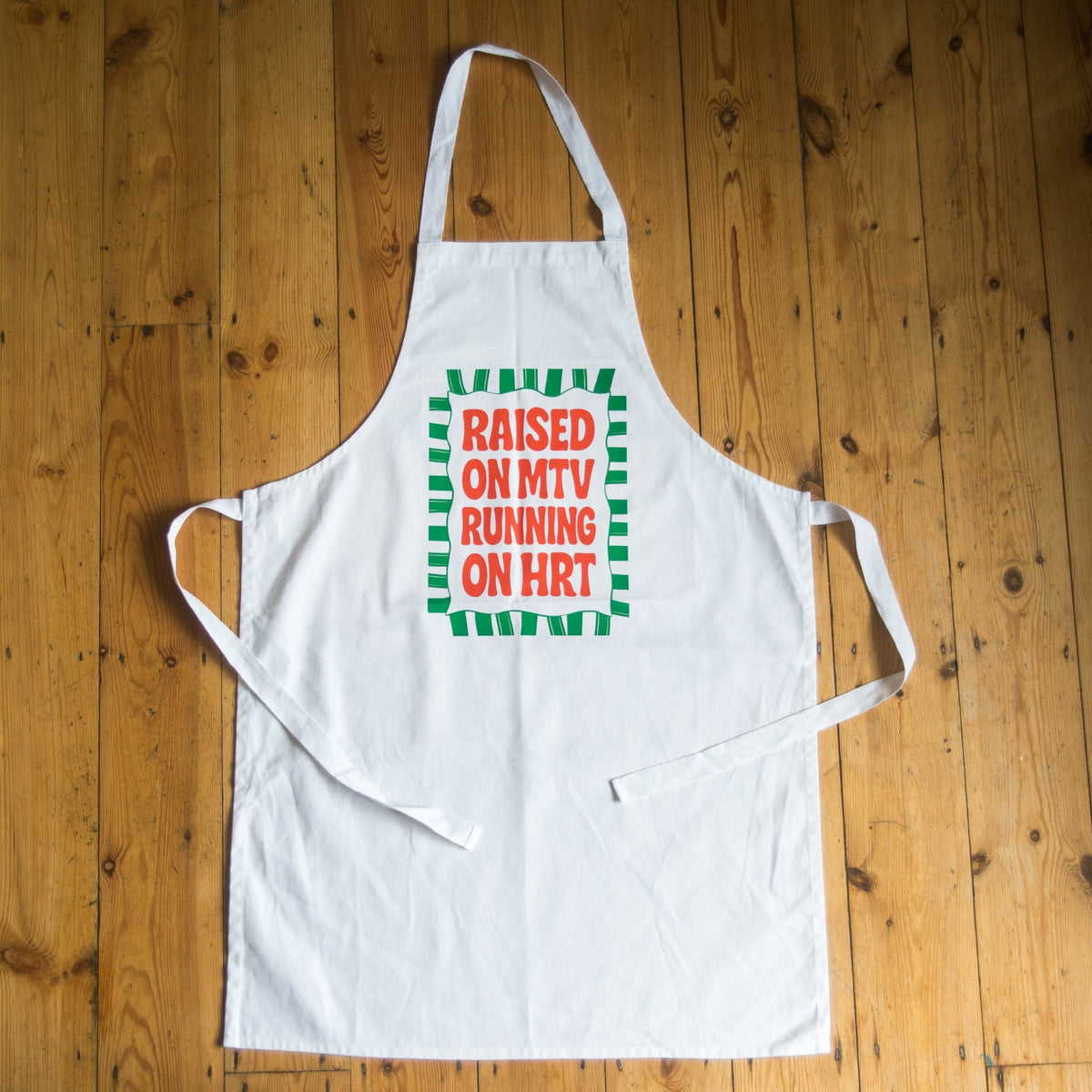 A white apron laid flat on a wooden plank surface, featuring a green and red graphic on the chest that reads 'RAISED ON MTV RUNNING ON HRT'.
