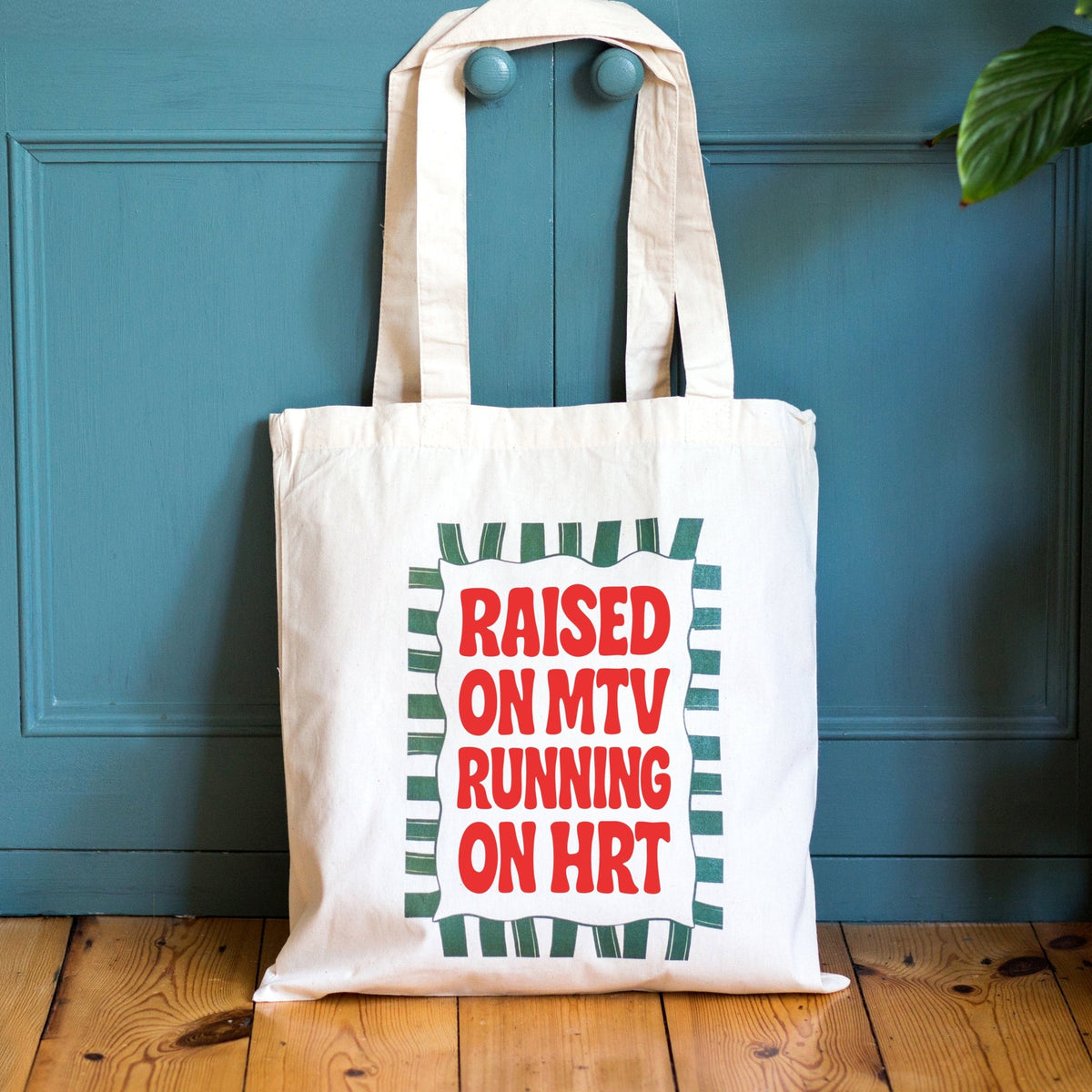 A cream canvas tote bag with long handles, featuring a graphic print that reads 'RAISED ON MTV RUNNING ON HRT' in red letters inside a white box with a green striped border. The bag is photographed leaning against a teal wooden door on a light wood floor.