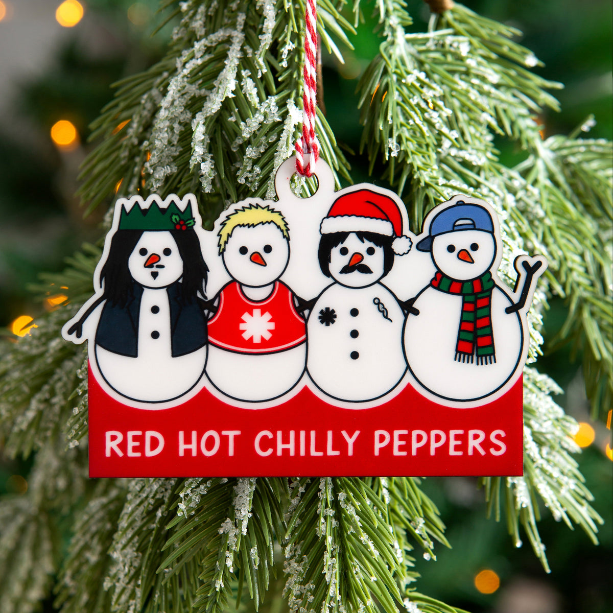 A Christmas ornament shaped like a red sign with four cartoon snowmen hanging from a flocked pine tree branch. The sign reads 'RED HOT CHILLY PEPPERS' in white text, and the background shows a blurred Christmas tree with warm lights.