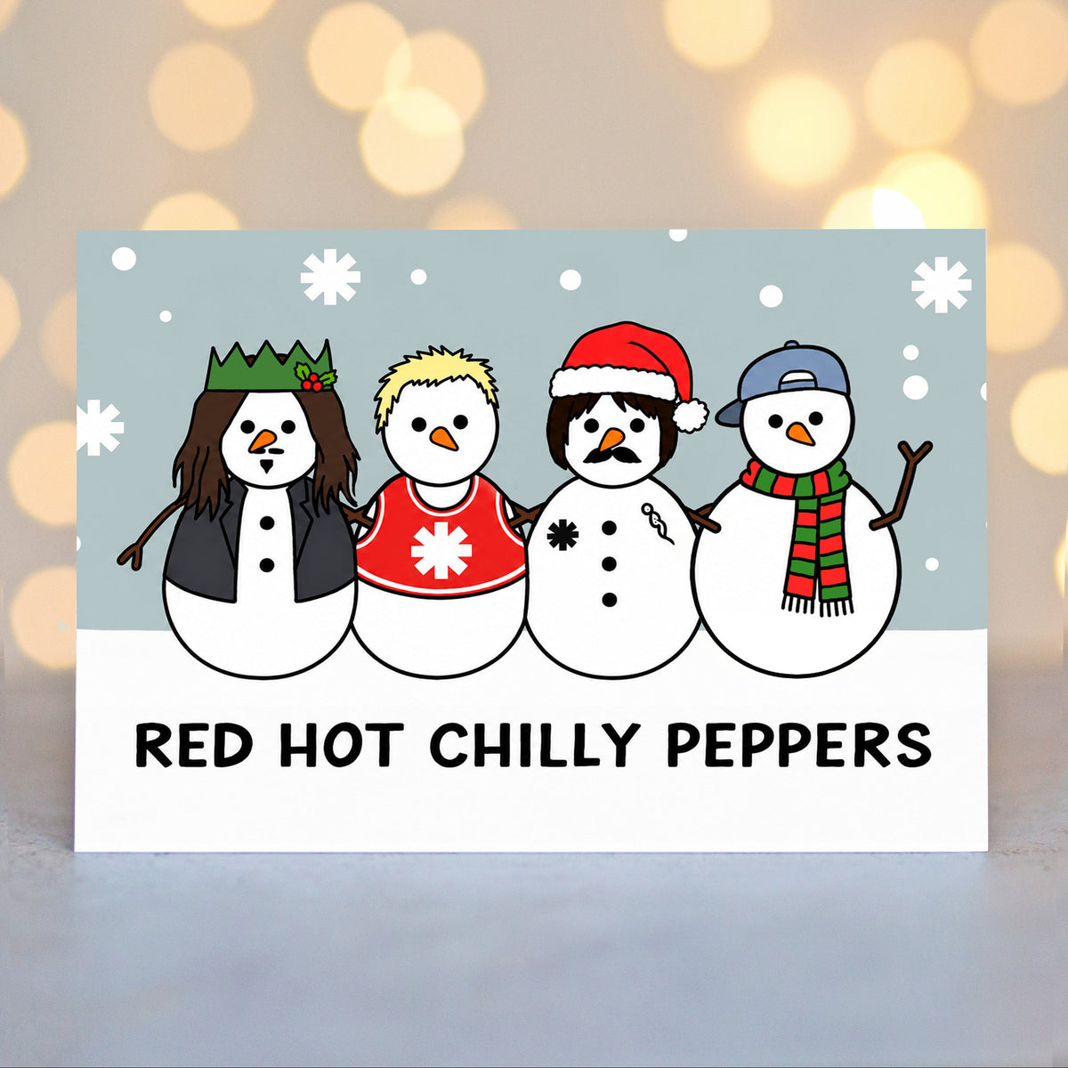 A Christmas card featuring a cartoon illustration of four snowmen wearing red hot chilli pepper themed hats and outfits, with the text 'RED HOT CHILLY PEPPERS' printed below. The card is displayed against a soft-focus background with warm golden bokeh lights.