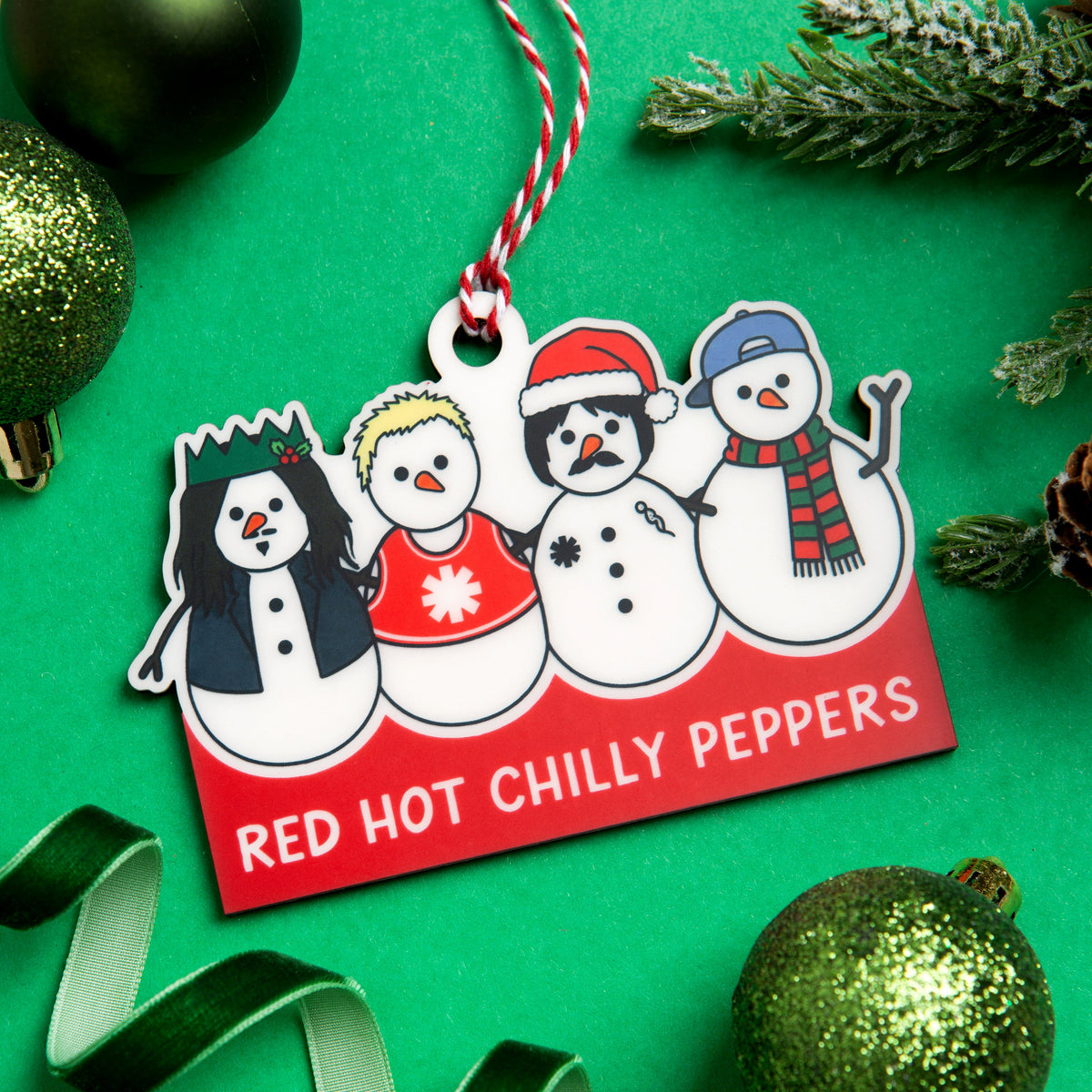 A festive Christmas ornament featuring four snowmen wearing winter hats and scarves, with the text 'RED HOT CHILLY PEPPERS' on a red banner. The ornament hangs from a red-and-white striped string and is photographed on a bright green background with holiday decorations including green ornaments, pine branches, and a green ribbon.