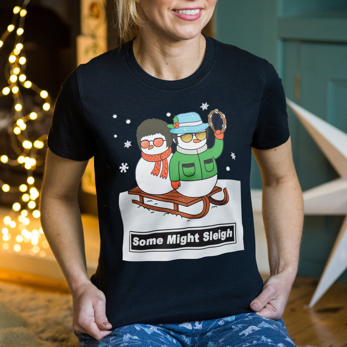 Person wearing a black Oasis themed Christmas t-shirt with a graphic of two snowmen and the text 'Some Might Sleigh' in a festive indoor setting.