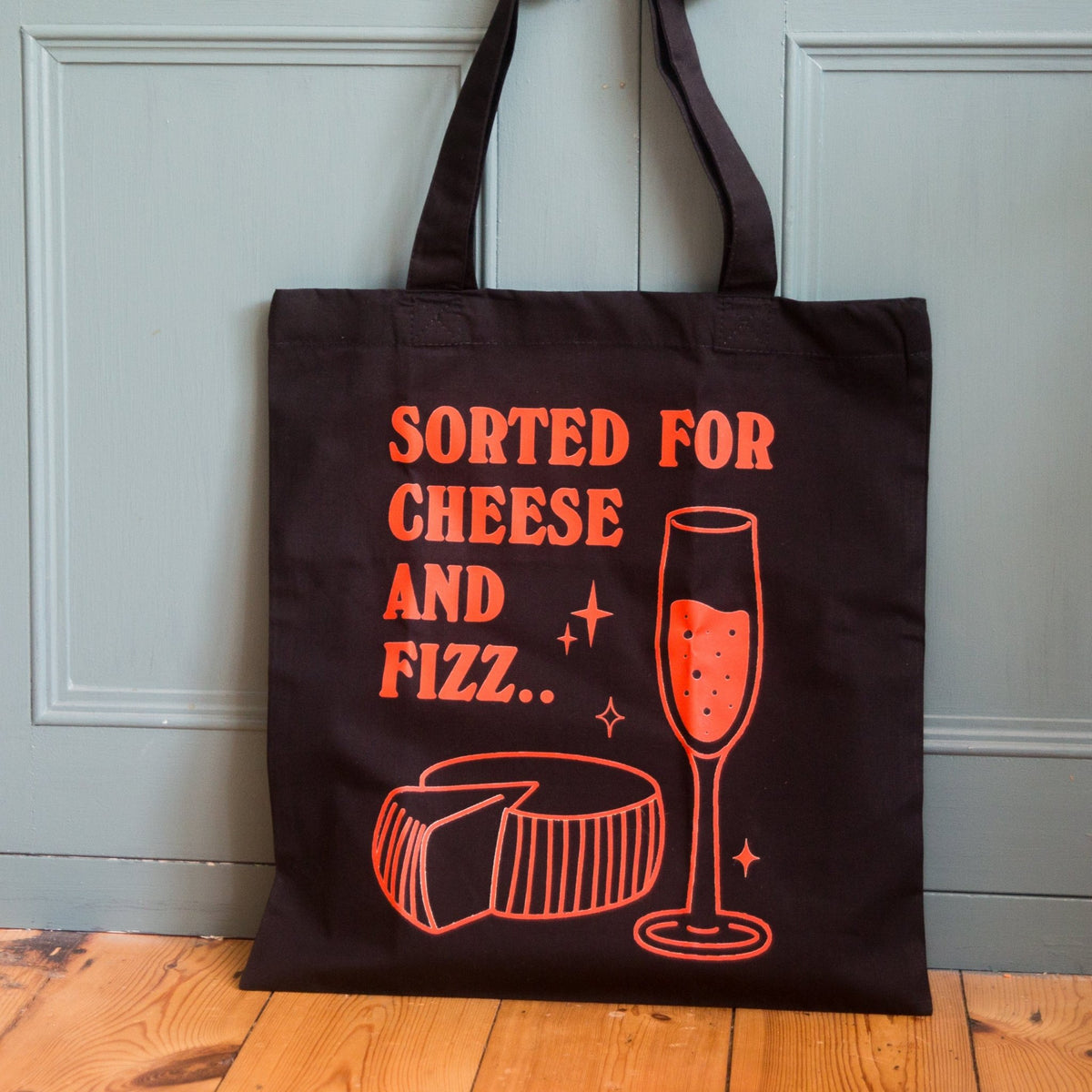 A black canvas tote bag with orange text reading 'SORTED FOR CHEESE AND FIZZ.