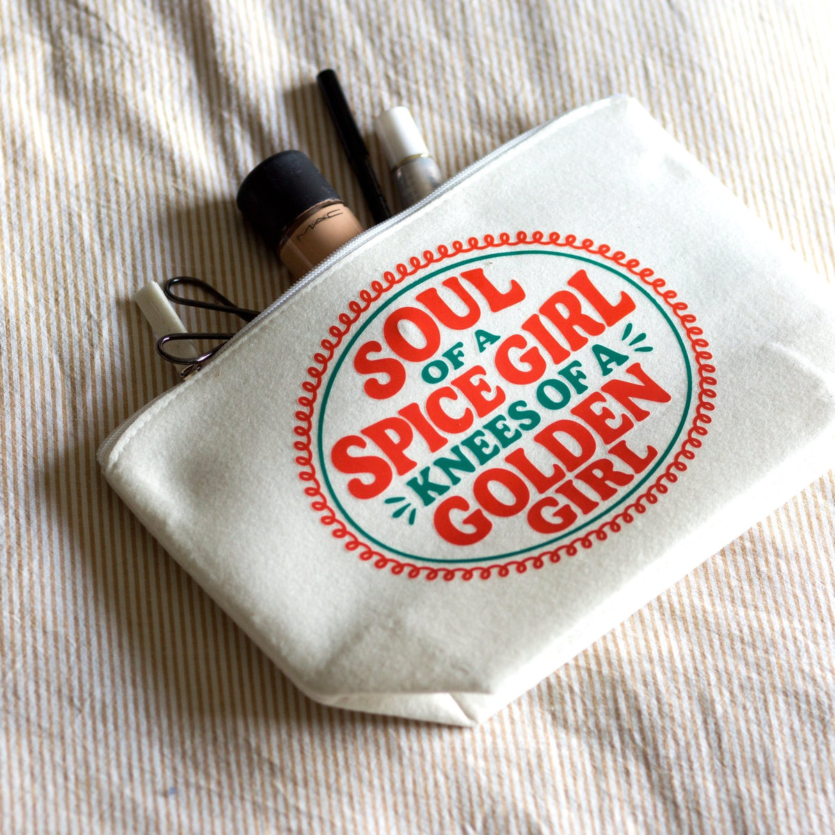 A white canvas cosmetic pouch with a red and green graphic reading 'SOUL OF A SPICE GIRL KNEES OF A GOLDEN GIRL,' photographed on a beige striped fabric background with makeup items peeking out from the top.