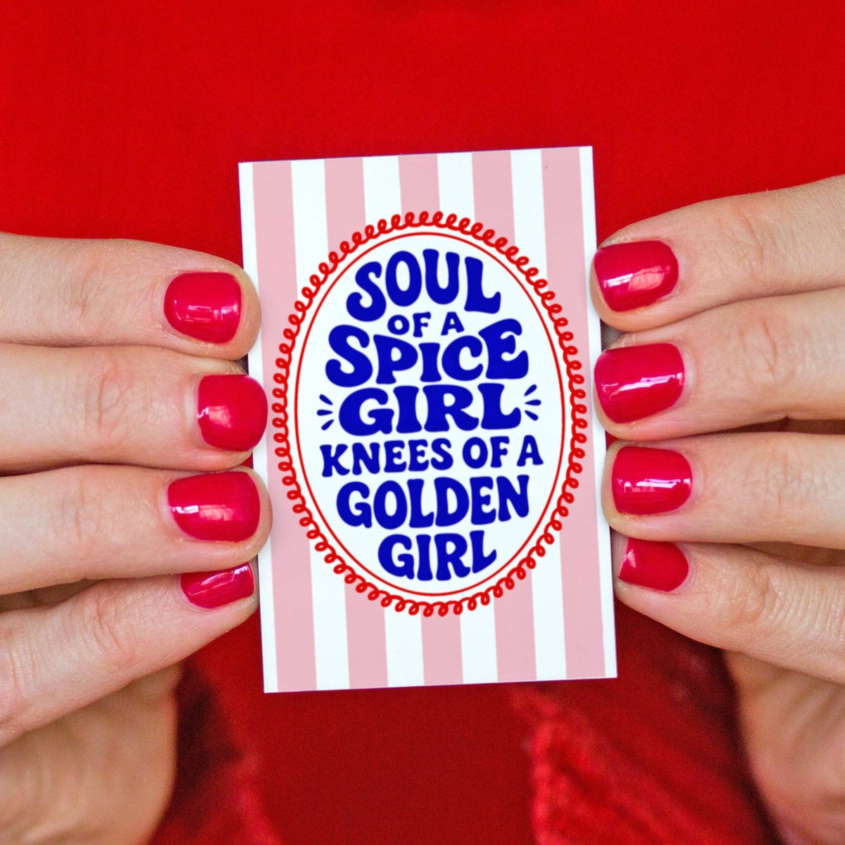 a fridge magnet with pink stripes and text in blue saying sould of a spice girl, knees of a golden girl held in a ladies hands against a red jumper