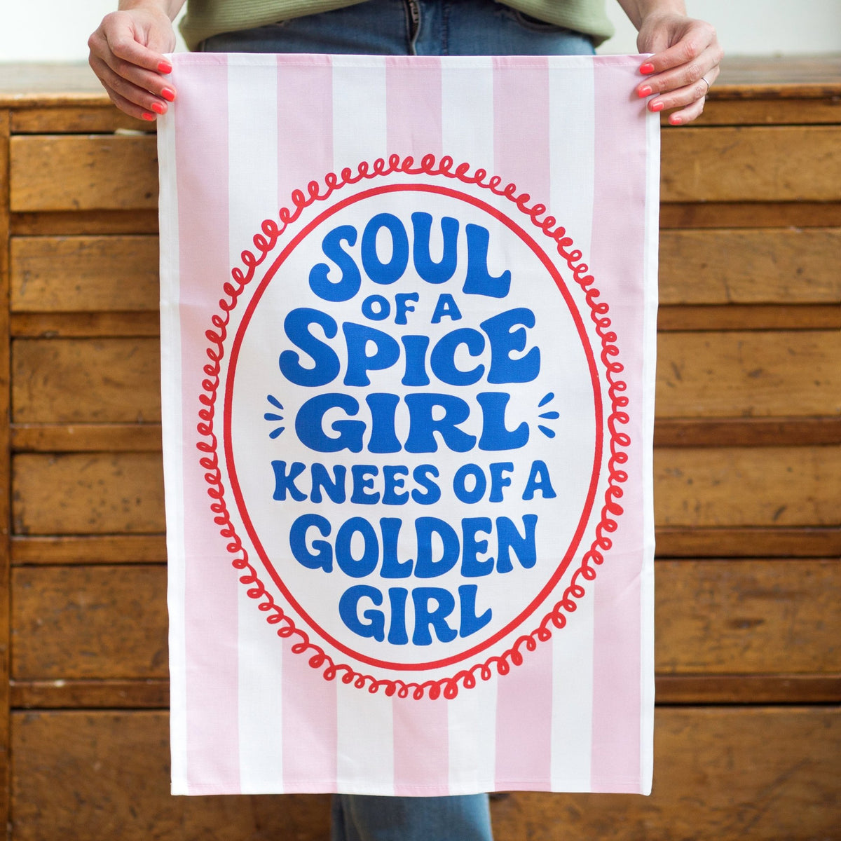 a striped tea towel with the slogan soul of a spice girls knees of a golden girl held against a wooden background