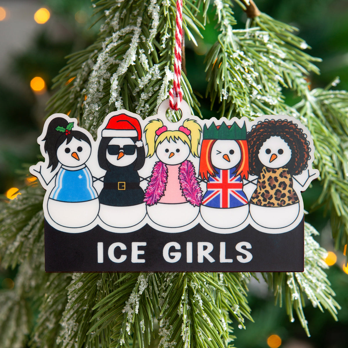 A Christmas ornament shaped like five cartoon snowmen dressed as The Spice Girls on a black bar, hanging from a red string on a frosted green Christmas tree with warm bokeh lights in the background. The pun 'ICE GIRLS' is printed on the ornament.