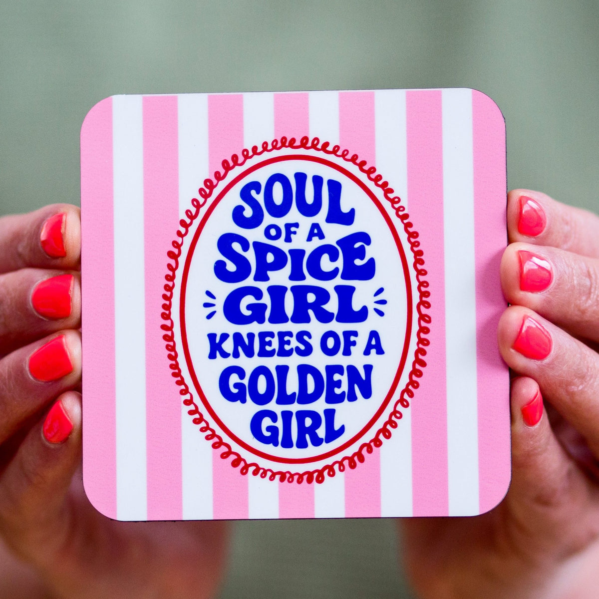Coaster with pink and white stripes and blue text held by hands with red nail polish.