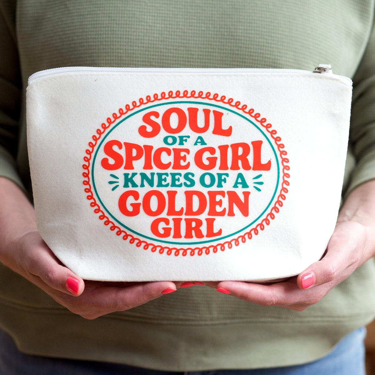 A person holding a white canvas zipper pouch with a colorful graphic that reads 'SOUL OF A SPICE GIRL KNEES OF A GOLDEN GIRL' in red and teal text.