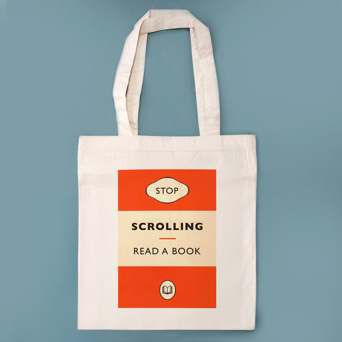 A cream canvas tote bag with a retro book cover design featuring orange-red blocks and the text 'STOP SCROLLING READ A BOOK' on a muted blue background.