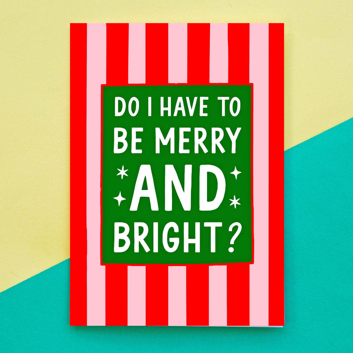 A Christmas card with red and pink vertical stripes and a green central panel reading “DO I HAVE TO BE MERRY AND BRIGHT?” in white text, photographed against a split cream and teal background.