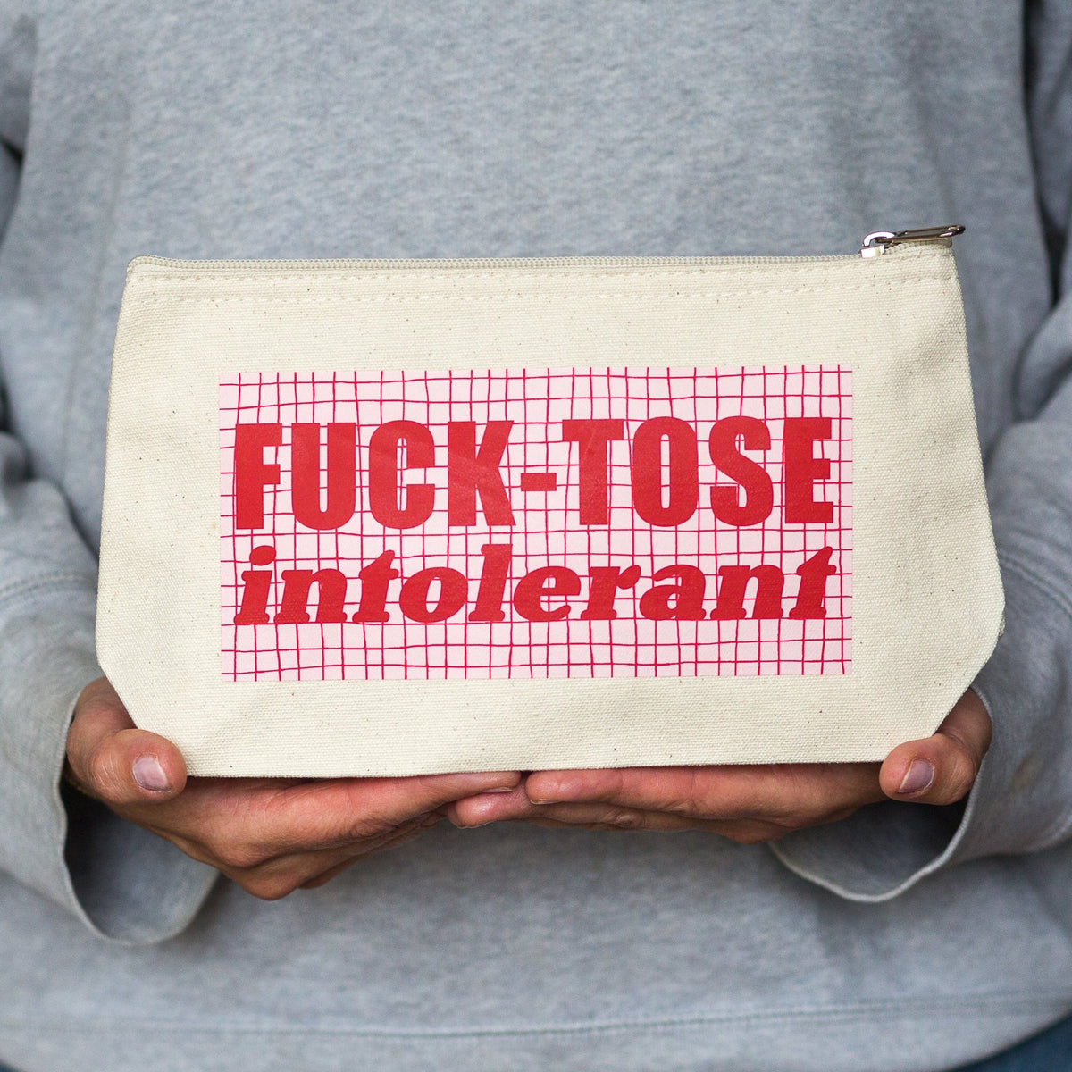 A person in a gray sweatshirt holds a cream canvas zipper pouch with red text reading 'FUCK-TOSE intolerant' over a red grid pattern.