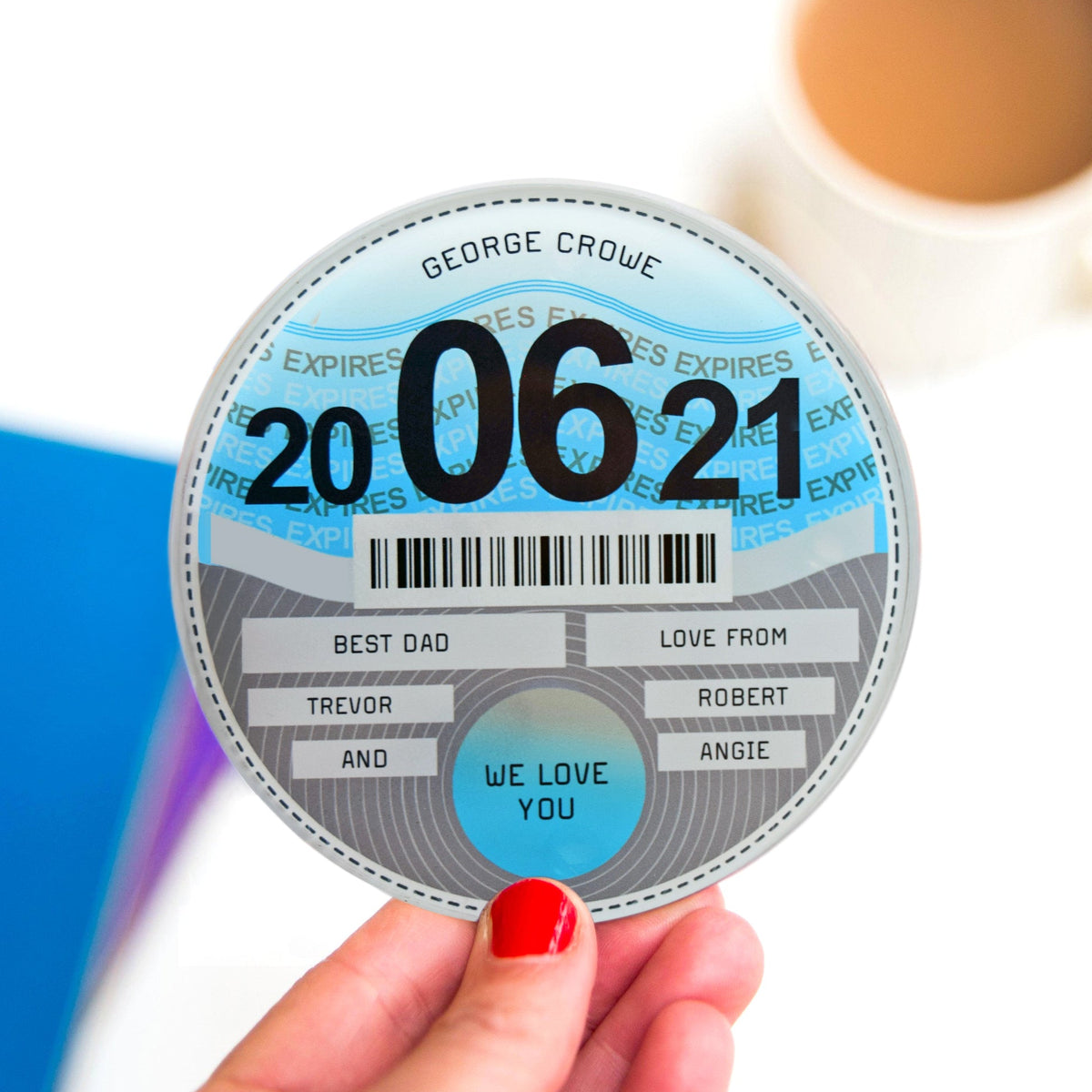A hand with red nail polish holds a round novelty expiration sticker that reads 'GEORGE CROWE' and '20 06 21', with decorative text and a barcode. A blurred coffee cup appears in the background on a white surface.