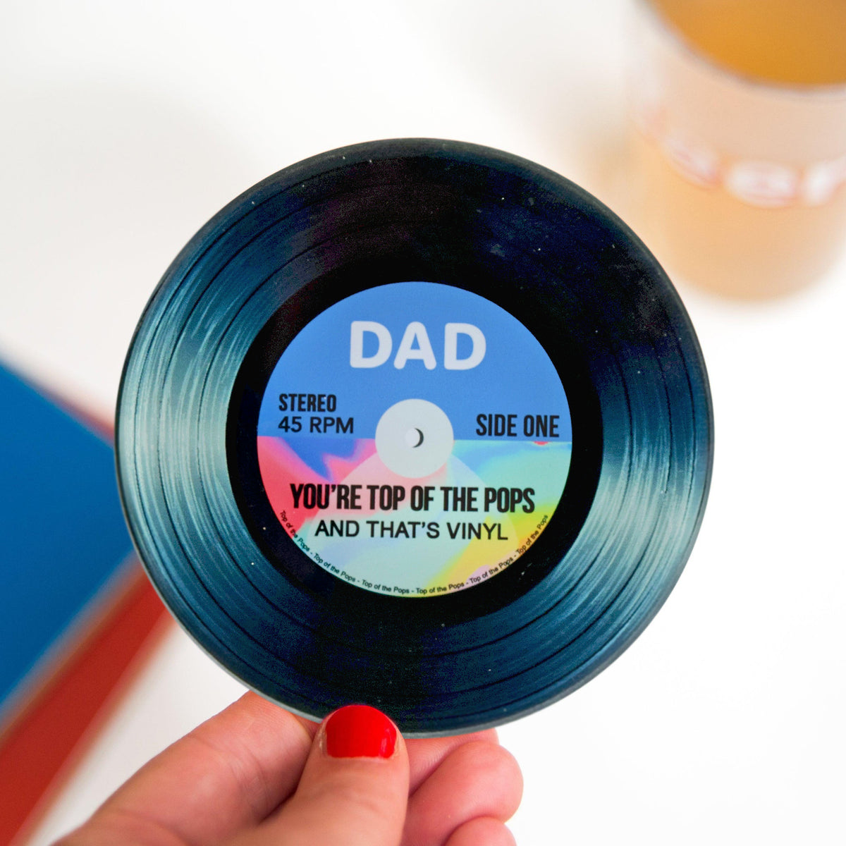 A hand with red nail polish holds a novelty record-shaped product labeled 'DAD' with the phrase 'YOU'RE TOP OF THE POPS AND THAT'S VINYL' against a softly blurred background.