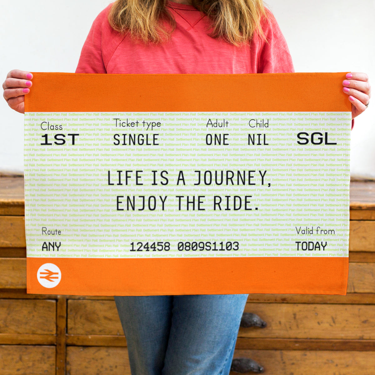 A person holding a large novelty train ticket tea towel featuring the words 'LIFE IS A JOURNEY, ENJOY THE RIDE.' and the British Rail logo, with wooden drawers in the background.