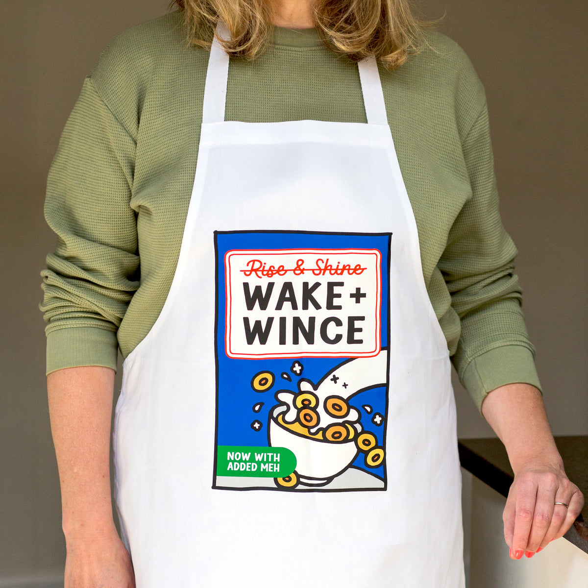 A person wearing a white apron with a colorful cereal box graphic that reads 'Riso & Shine WAKE + WINCE' and 'NOW WITH ADDED MUSH', over an olive green long-sleeve top.