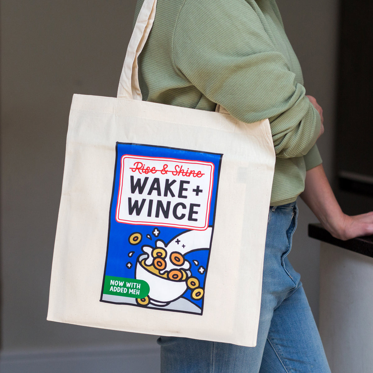 A person wearing a sage green ribbed sweater and blue jeans carries a cream canvas tote bag. The bag features a parody graphic of a cereal box labeled 'Rise & Shine WAKE + WINCE' with blue, red, yellow, and green details.