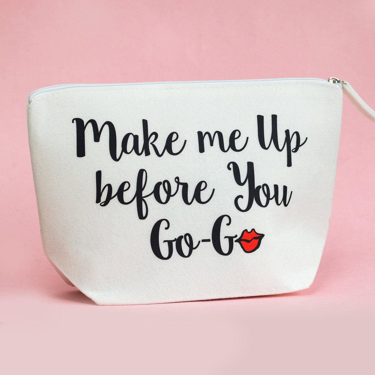 An off-white cosmetic pouch with the phrase 'Make me Up before You Go-Go' in black script, accented by a small red lips graphic, set against a solid pink background.